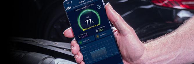 Das Bild zeigt ein Smartphone in der Hand vor einer Batterie. Darauf wird der Ladezustand in der Battery-Guard App angezeigt.