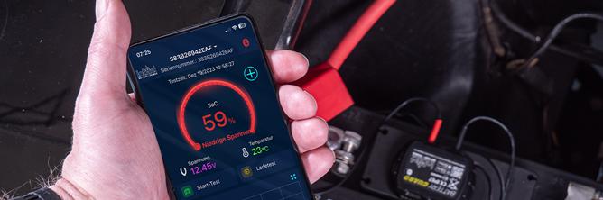 Das Bild zeigt ein Smartphone in der Hand vor einer Batterie. Darauf wird der Batterie-Ladezustand in der Battery-Guard App kontrolliert.