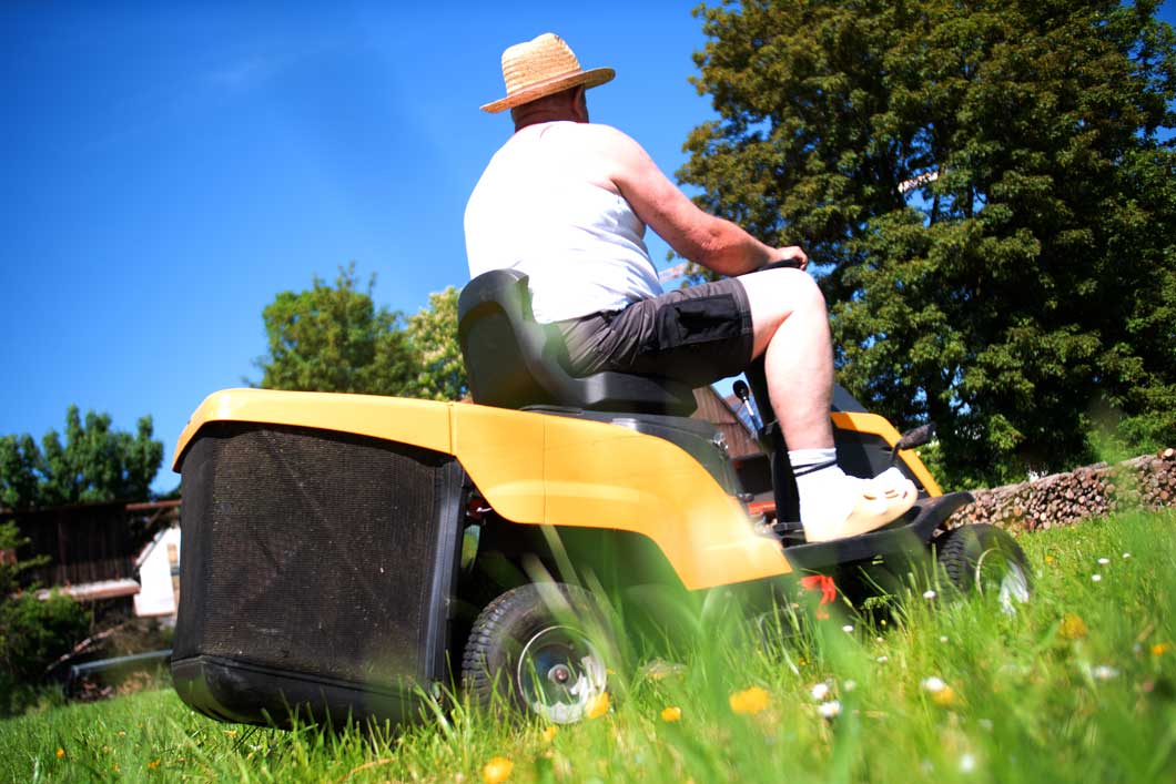 Teaserbild Einsatzgebiet Rasentraktor-Batterien Das Bild zeigt einen Mann mit Strohhut, welcher gerade auf seinem gelben Aufsitzrasenmäher bei Sonnenschein den Rasen mäht.